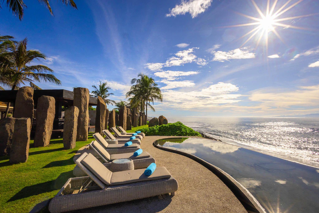 Oceanfront infinity pool with sun loungers under a bright tropical sun, surrounded by palm trees and stone pillars—an ideal beachfront escape for wellness retreats in Bali.