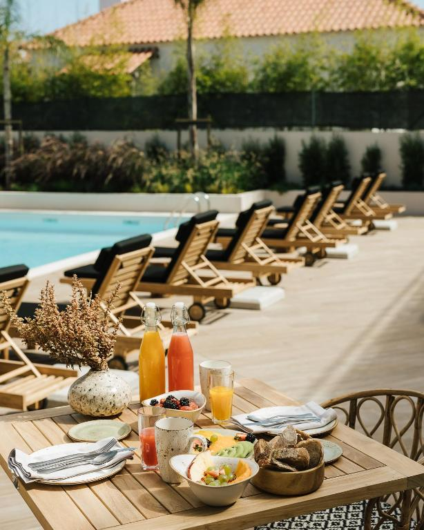 Elegant outdoor breakfast setup on a wooden table, featuring fresh fruit bowls, bread, and colorful juices in glass bottles, with a serene poolside view of wooden lounge chairs and lush greenery in the background