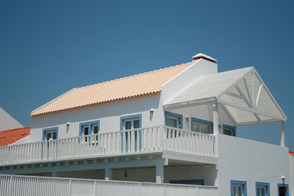 Charming white house with a terracotta-tiled roof, blue window frames, and a spacious balcony featuring a decorative pergola, set against a clear blue sky.