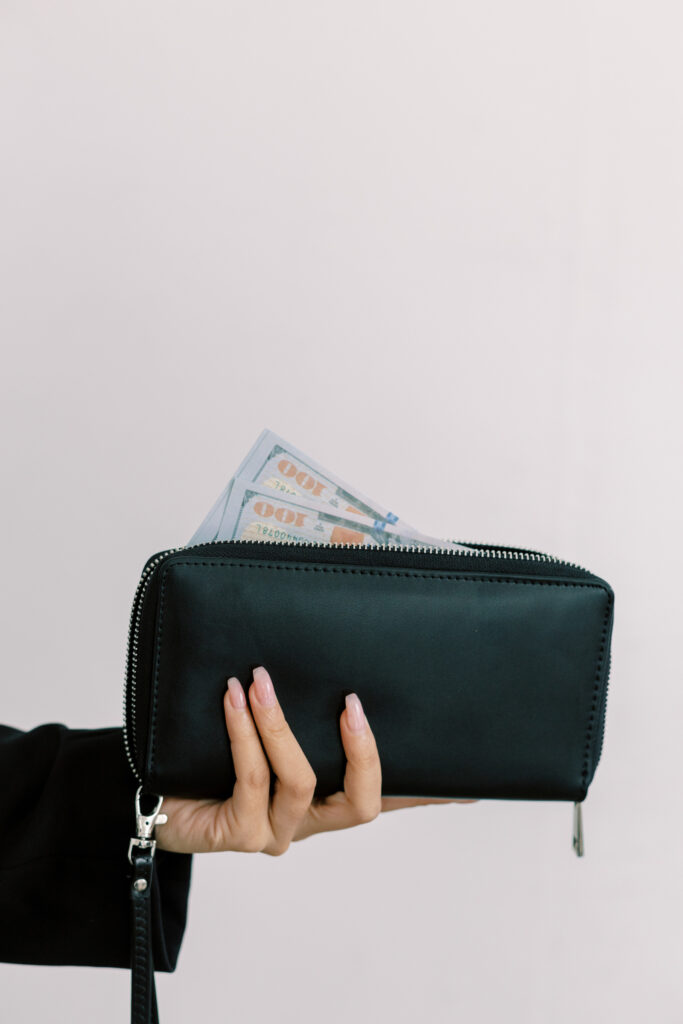 A stylish hand with manicured nails holds a sleek black wallet, with crisp hundred-dollar bills peeking out, symbolizing financial success, abundance, and wealth. The minimalist background keeps the focus on the wallet and cash, evoking themes of entrepreneurship, smart money management, and financial freedom.
