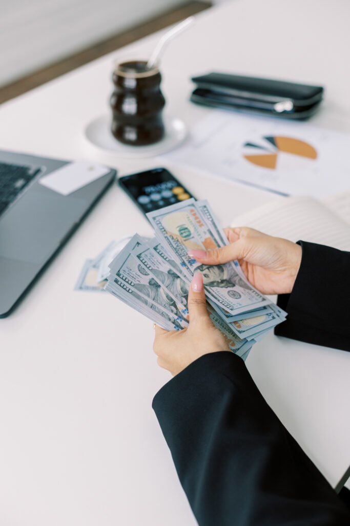 A woman in a black blazer counting stacks of $100 bills at a modern, well-organized workspace. A laptop, smartphone with a calculator app open, financial documents with pie charts, and a glass of iced coffee are on the desk, symbolizing profits, entrepreneurship, and business growth.