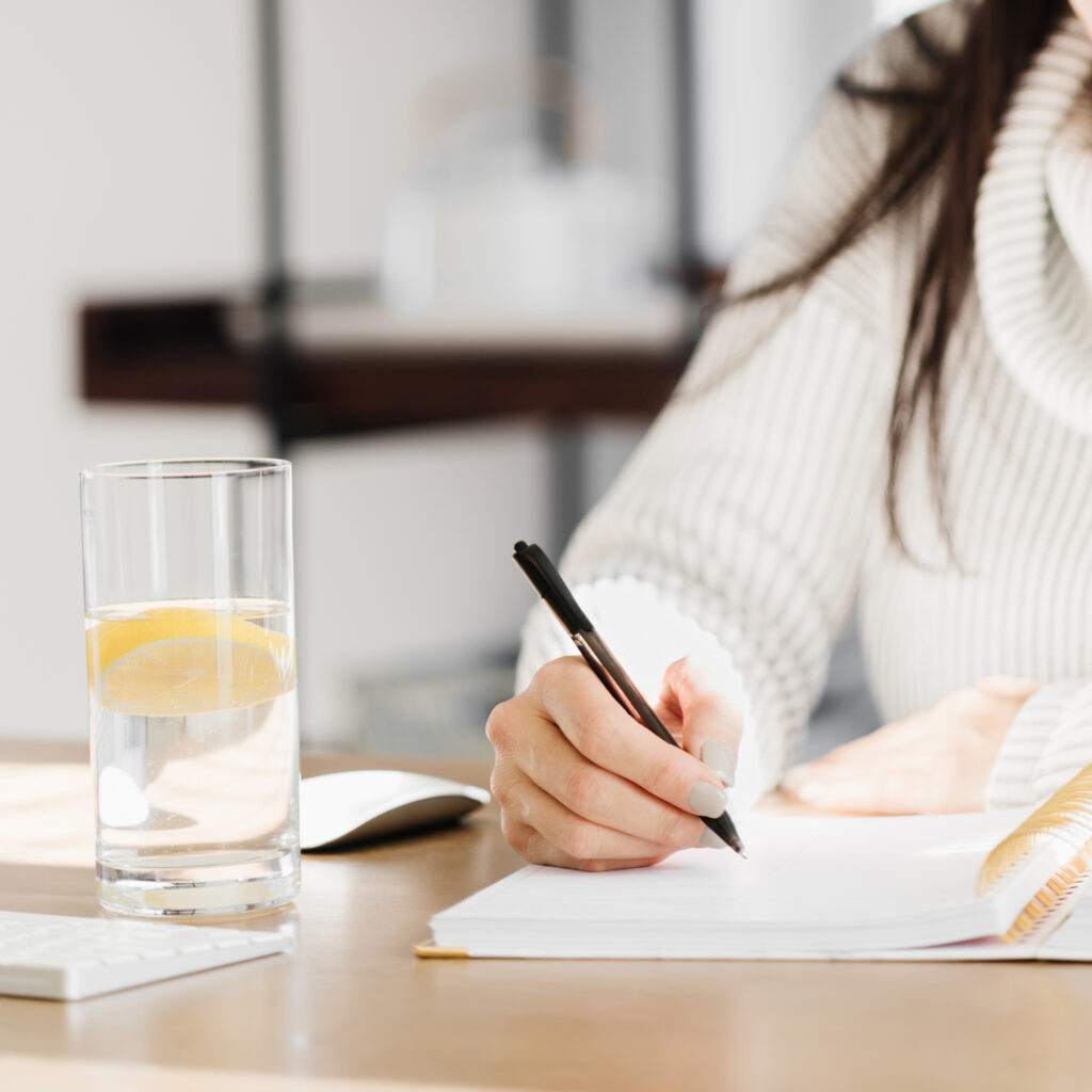 Person in cream sweater writing in notebook with pen next to glass of lemon water on wooden desk