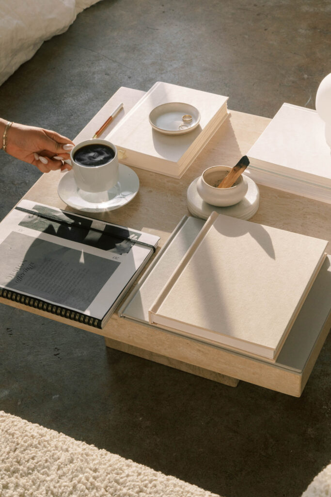 Minimal coffee table scene in warm sunlight with stacked neutral colored books and notebooks. A hand holds a cup of black coffee beside a small dish with two rings, a pen, a journal that reads “artists offerings,” and a ceramic holder with a burning incense stick.
