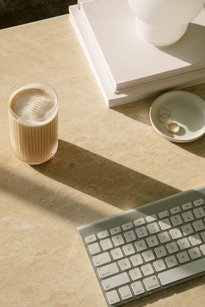 Sunlit stone desk with a glass of iced coffee casting a long shadow beside a wireless keyboard. Stacked white books and a small dish holding three gold rings sit nearby, creating a calm minimalist workspace scene.