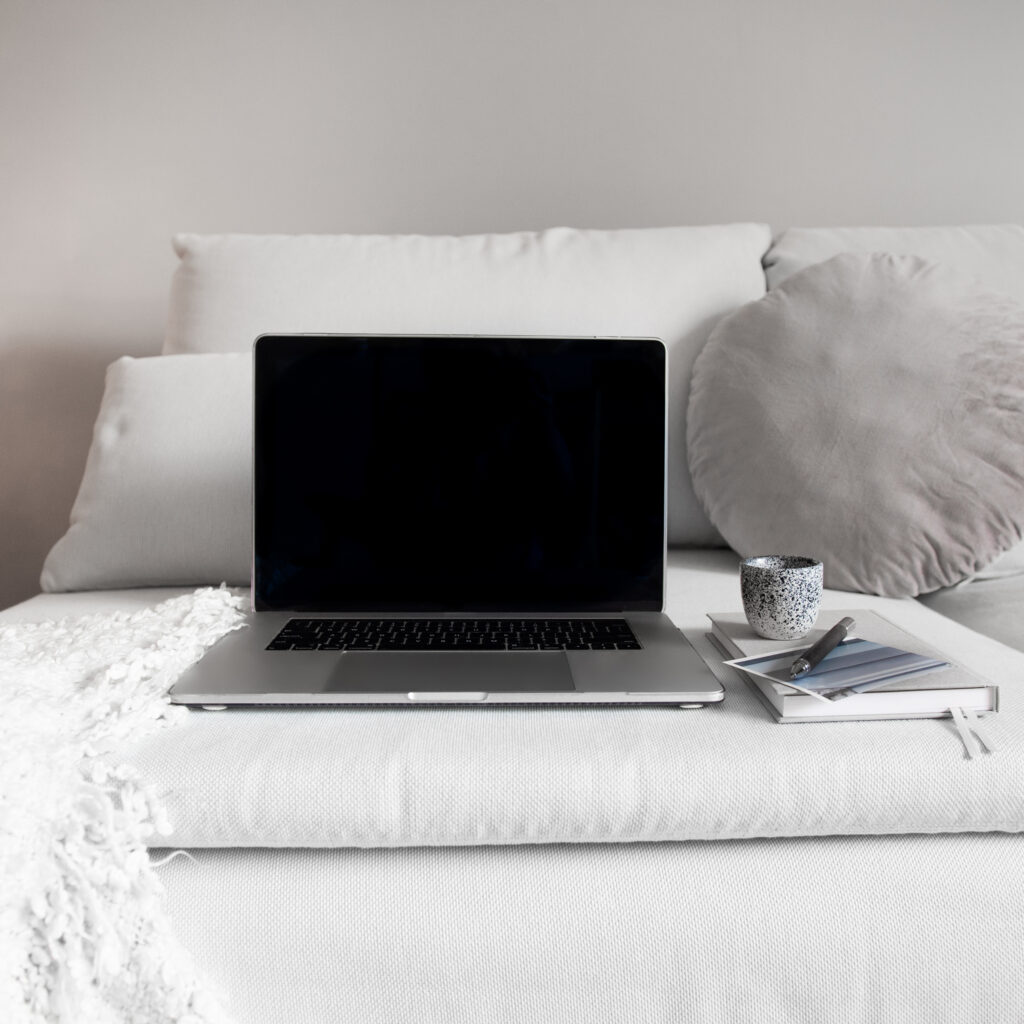 Laptop with black screen on white bed next to coffee cup and books in minimalist bedroom workspace setup