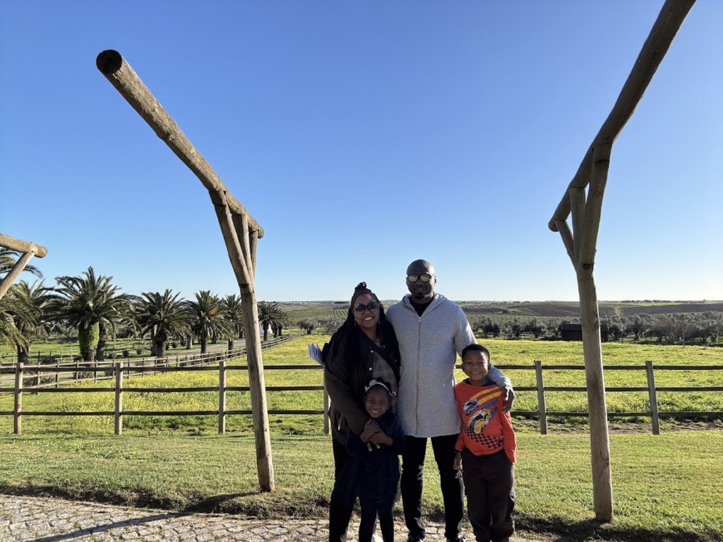 Family of four posing together at scenic farm with wooden fence, palm trees, and green pastures under blue sky