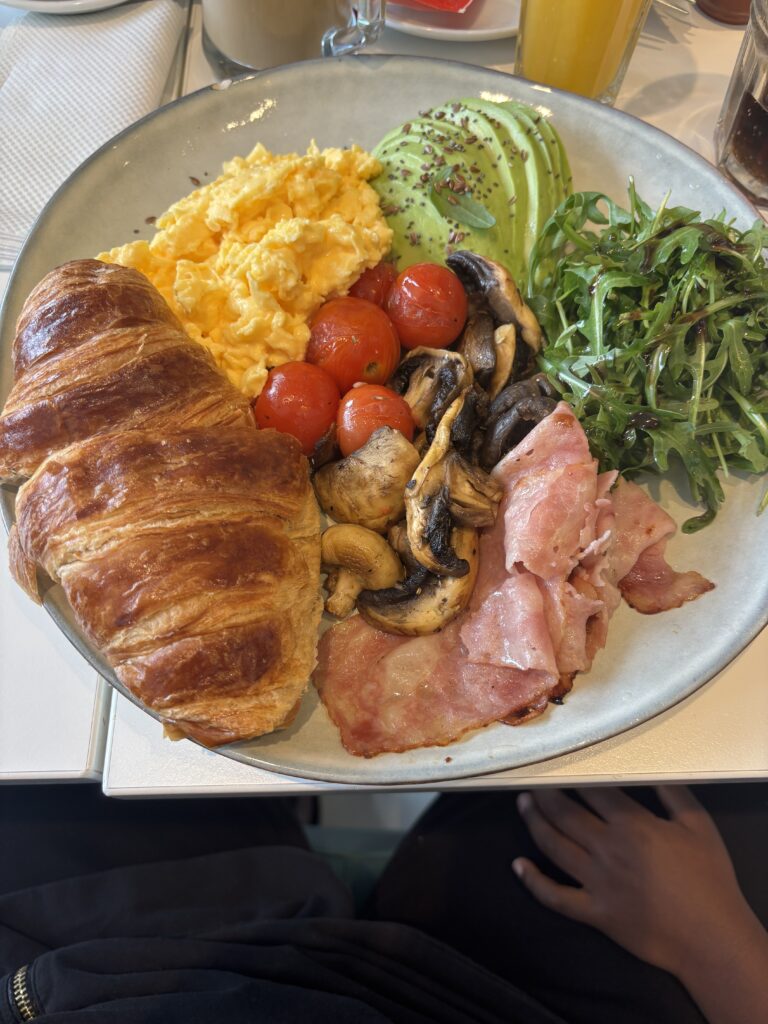 Breakfast plate with croissant, scrambled eggs, bacon, mushrooms, cherry tomatoes, avocado slices and arugula salad
