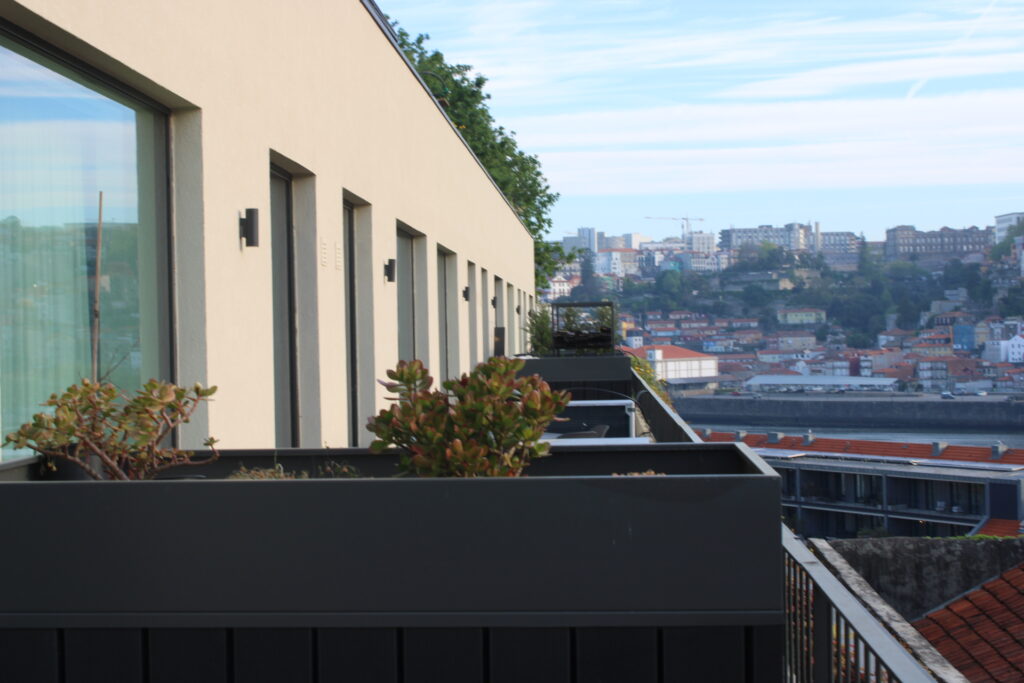 Modern beige building with narrow windows and rooftop garden planters overlooking cityscape with red tile roofs