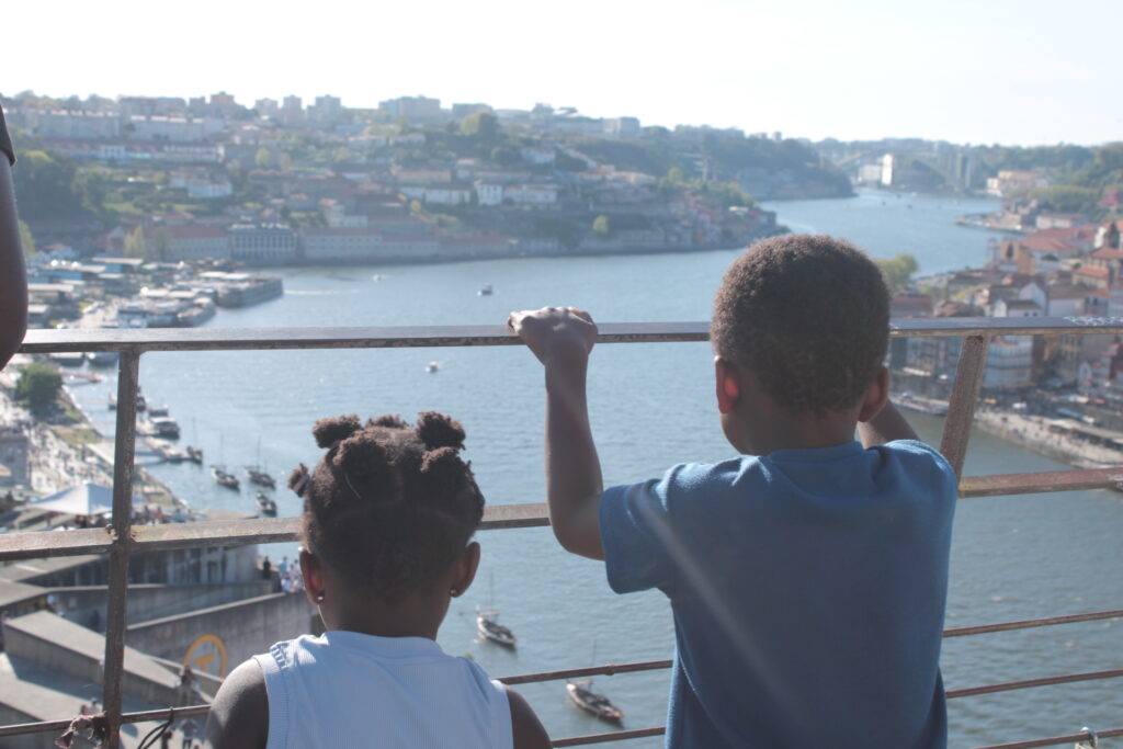 Two children looking out over a scenic river view with boats and hillside buildings from an observation deck railing