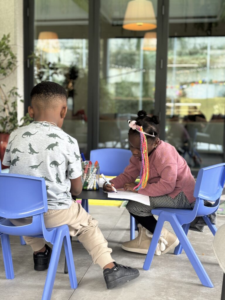 Two children sitting at blue plastic chairs drawing together at a table in a bright indoor space with large windows.