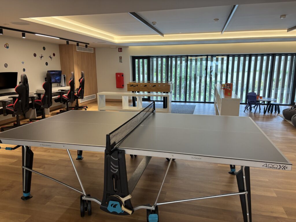 Modern game room with ping pong table, gaming chairs, foosball table, and large windows with vertical blinds