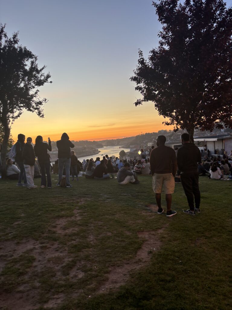 People gathering at sunset viewpoint overlooking river valley with orange sky and silhouetted trees in scenic park setting