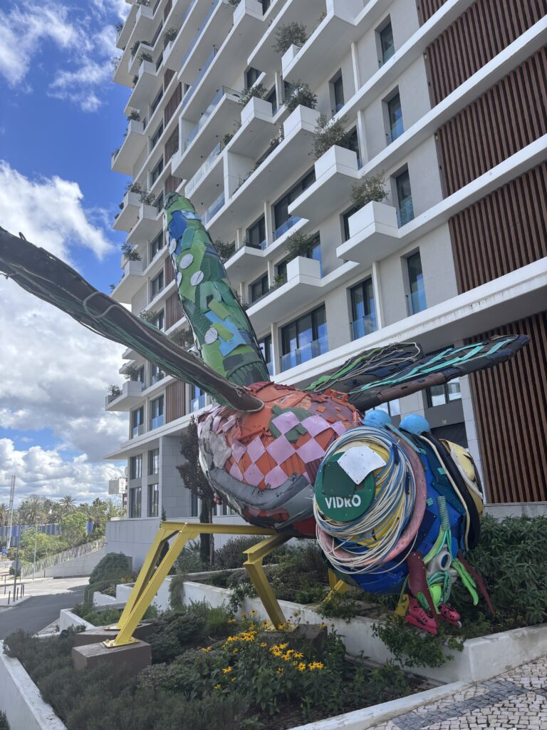 Large colorful hummingbird sculpture with mosaic patterns displayed outside modern white apartment building with balconies