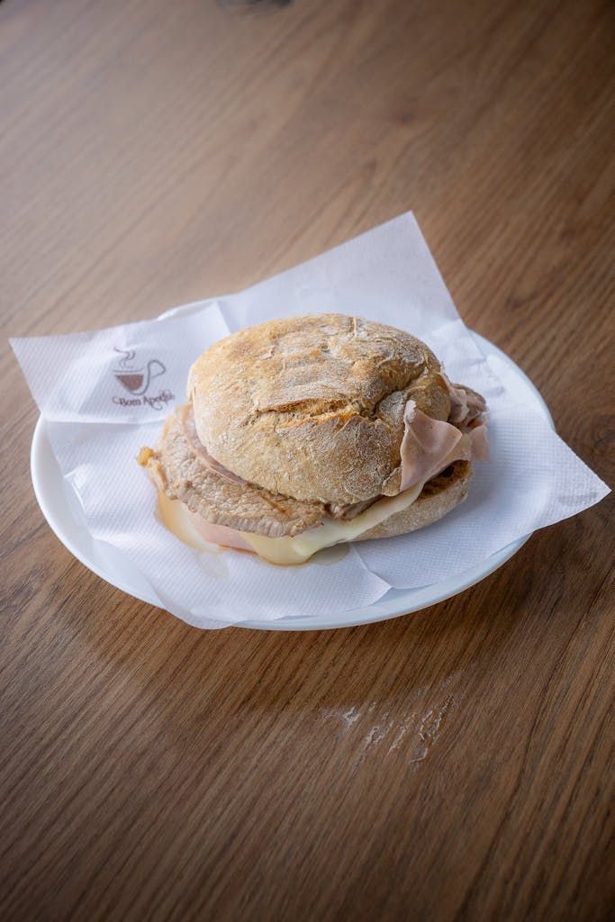 A savory bifana sandwich on a white plate with a rustic wooden table background.