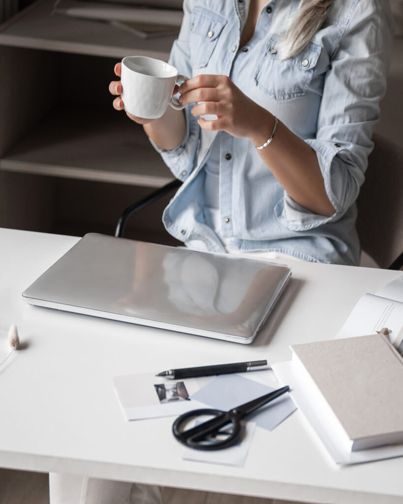 Woman in blue shirt drinking coffee while working on laptop at white desk with office supplies