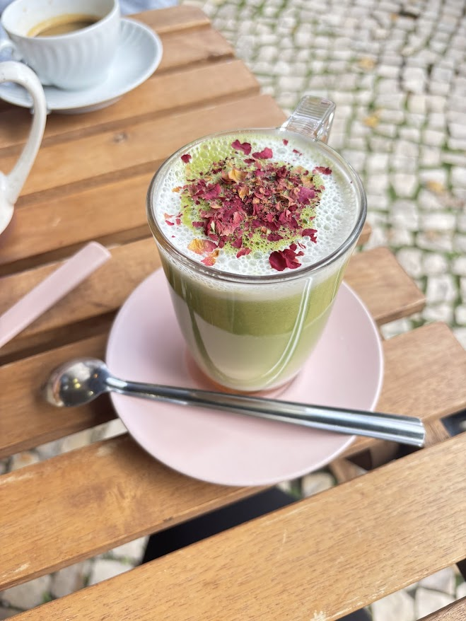 Matcha latte with rose petals and pistachios on pink saucer with spoon at outdoor cafe table