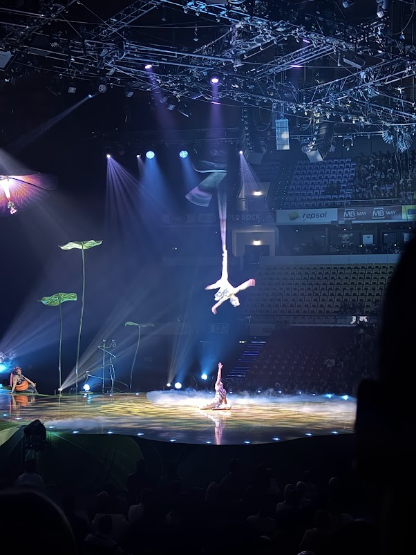Aerial acrobat performing suspended trapeze act under blue spotlights in circus arena with audience seating visible.