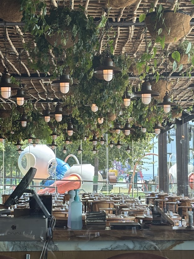 Modern restaurant interior with cascading green ivy, hanging pendant lights, marble counter, and contemporary art installations by windows.