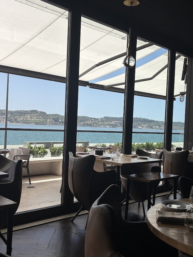 Waterfront restaurant with panoramic sea views, modern dining tables, floor-to-ceiling windows, and retractable awning.