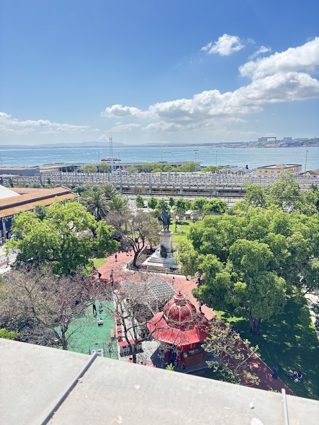 Aerial view of waterfront park with red pavilion, trees, statue, and bay cityscape under blue sky with clouds