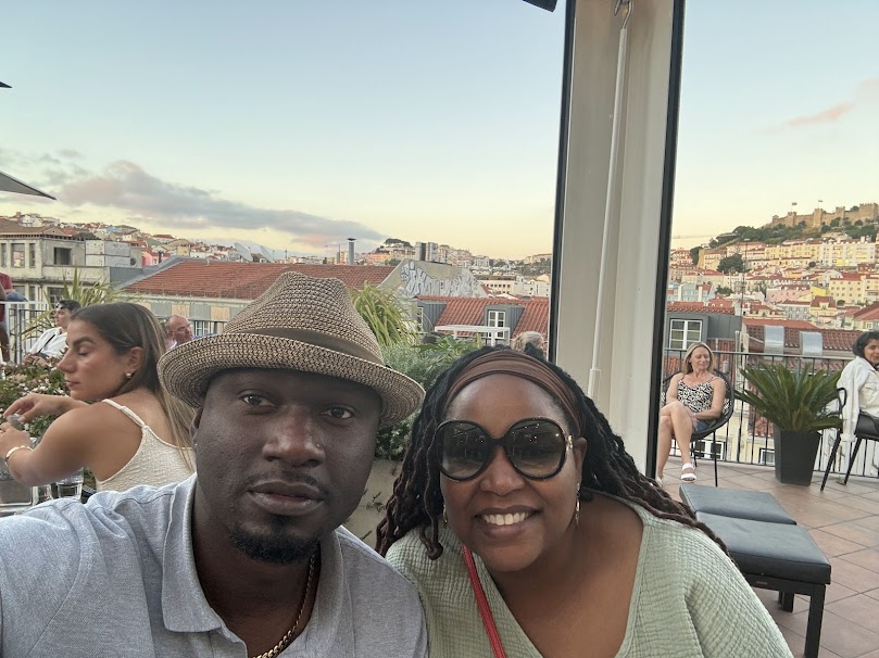 Two people smiling at a rooftop bar with Lisbon cityscape and hills in background at sunset.