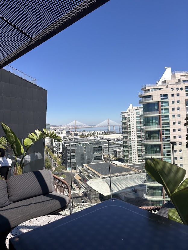 Rooftop terrace overlooking San Francisco Bay Bridge with modern buildings, plants, and seating area on clear blue sky day