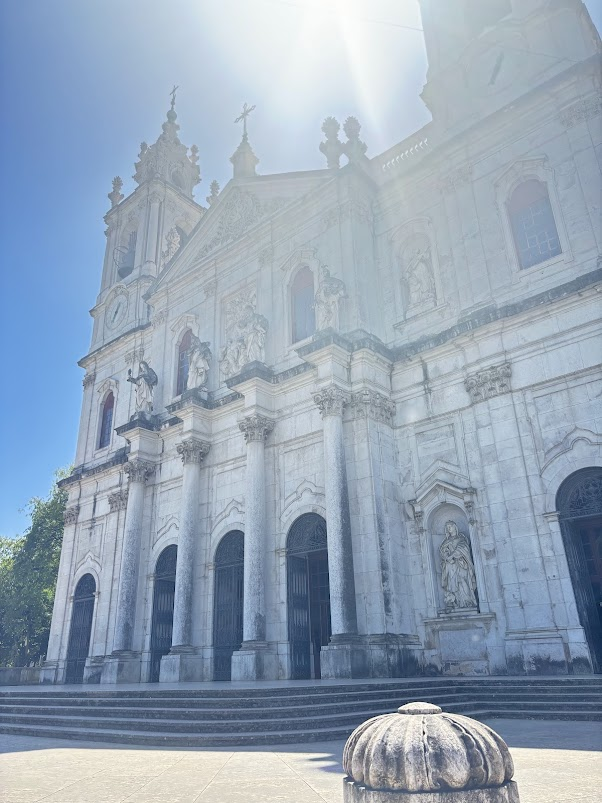 White baroque cathedral with ornate facade, twin towers with crosses, arched windows, and stone steps under bright sunlight.