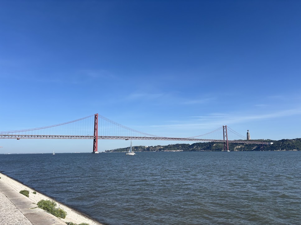 Red suspension bridge spanning blue water with sailboat and green hills under clear sky