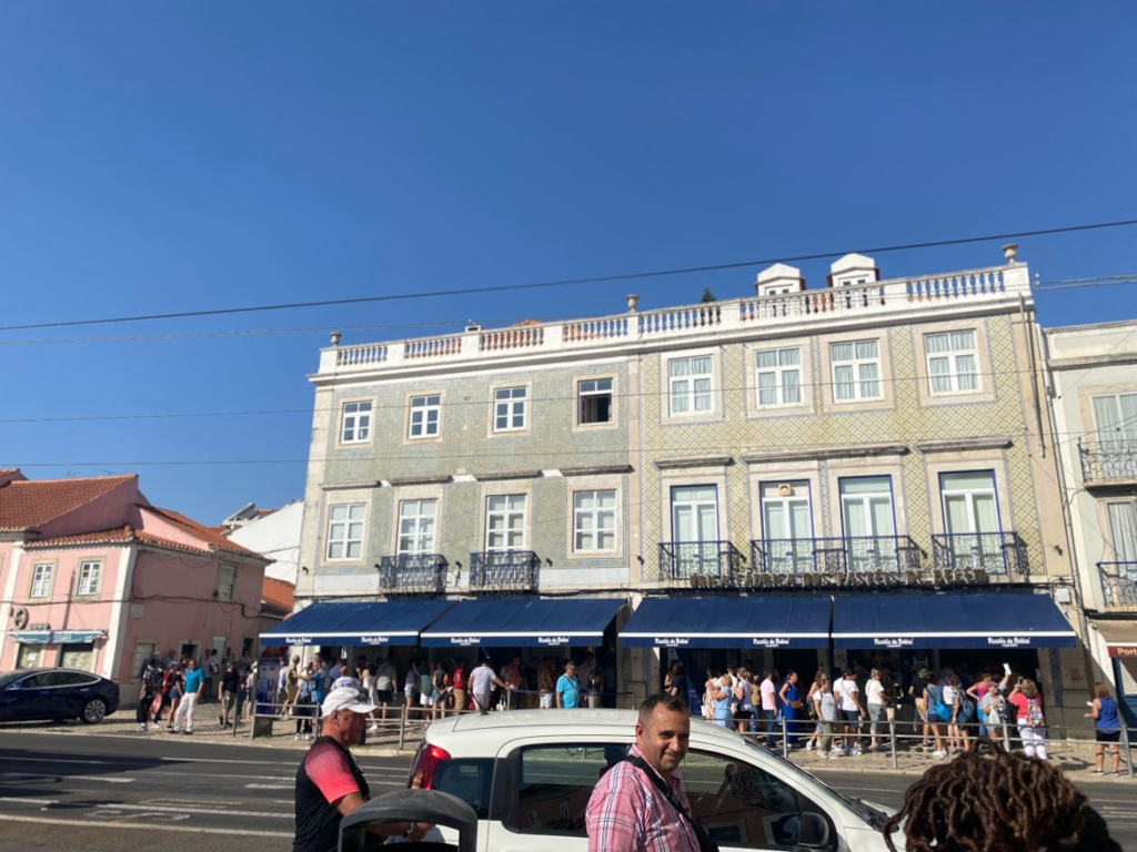 Charming European street scene with elegant cream-colored building, blue storefront, and crowds of visitors under clear sky.