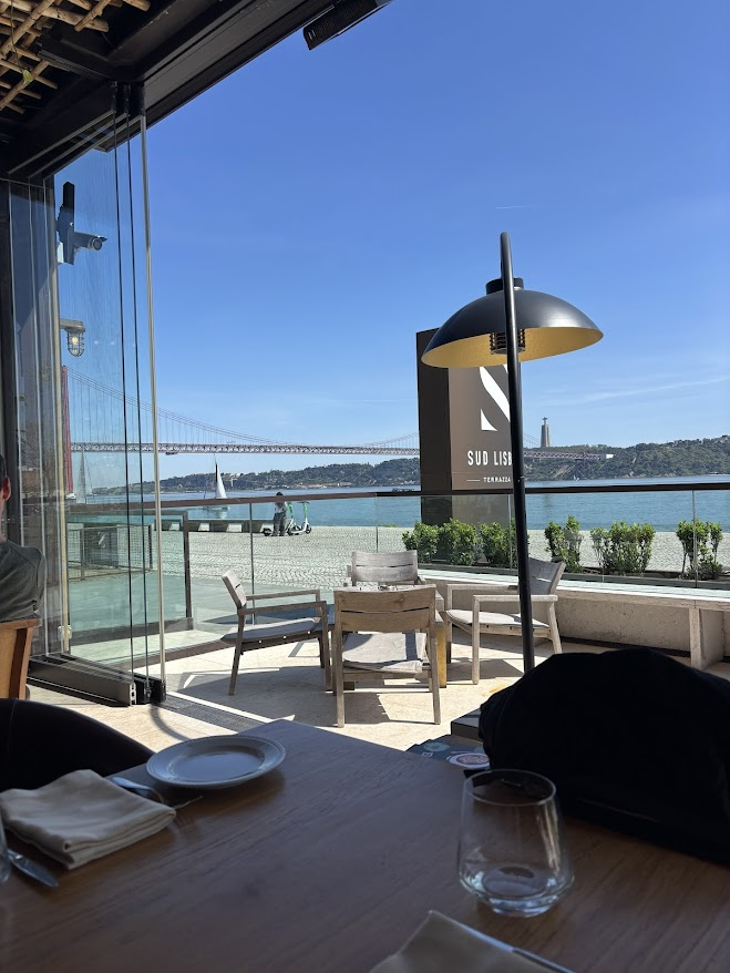 Upscale waterfront restaurant terrace with Golden Gate Bridge view, modern heating lamp, and bay seating area under clear blue sky.