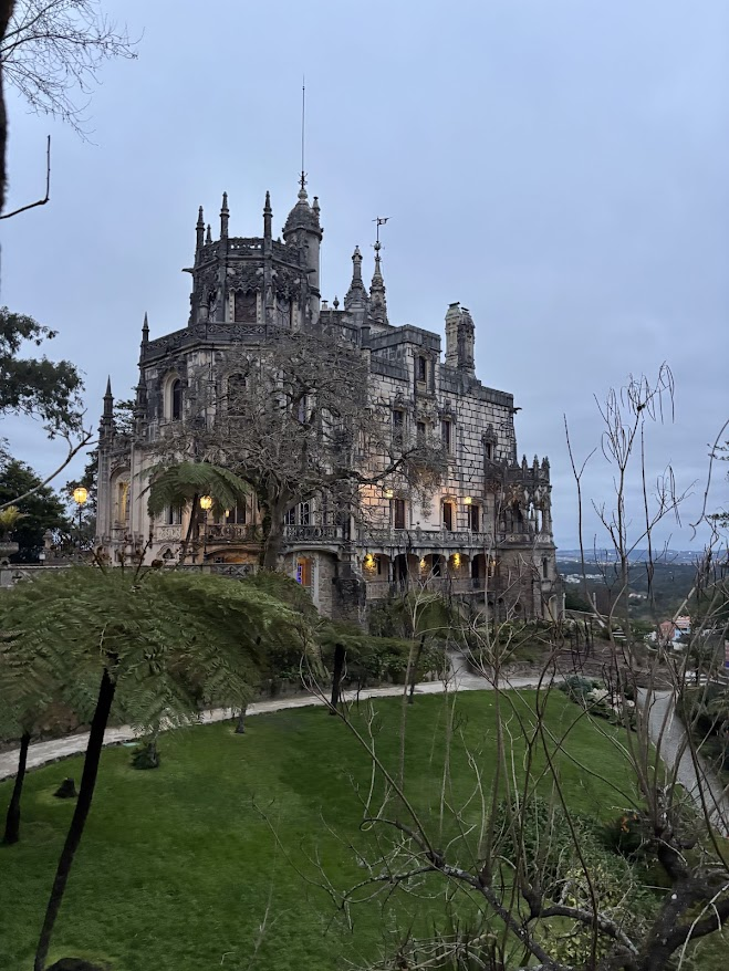 Gothic stone castle with multiple spires and turrets perched on a hilltop, surrounded by manicured gardens and bare trees.