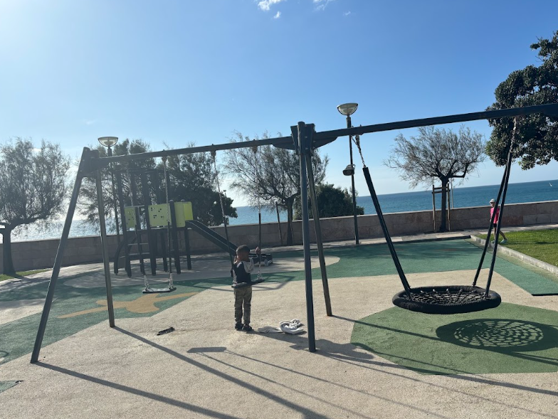 Child playing on modern playground with swings and climbing equipment overlooking ocean beach on sunny day.